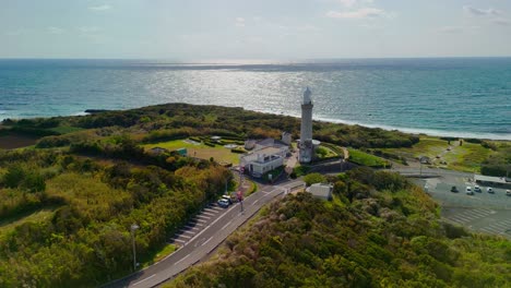 a stunning, static wide aerial shot captures a lighthouse on a lush green peninsula in japan, with the sun reflecting brightly on the vast ocean in the background