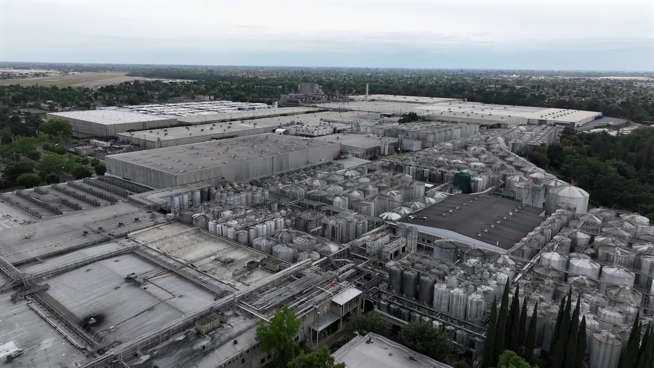 Impianto di lavorazione industriale ed edifici di magazzino presso la Gallo Winery a Modesto, California, con alberi verdi lussureggianti in primo piano sotto un cielo coperto negli Stati Uniti. Ripresa aerea con drone in avvicinamento.