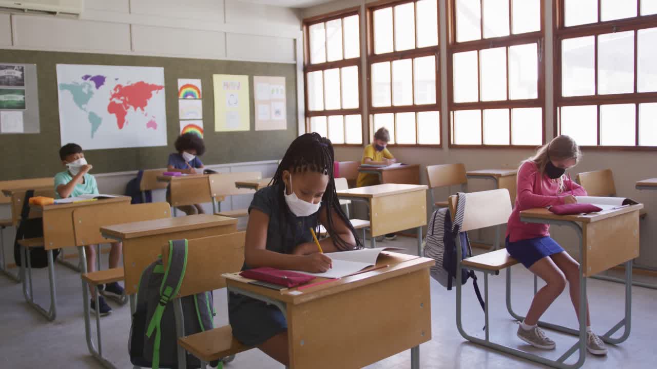 Grupo de crianças usando máscaras escrevendo enquanto estão sentadas em suas carteiras na escola.