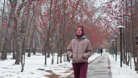 joven con chaqueta cálida y capucha camina por un camino nevado bordeado de árboles de bayas rojas, mirando a su alrededor con curiosidad, distante figura borrosa en el fondo