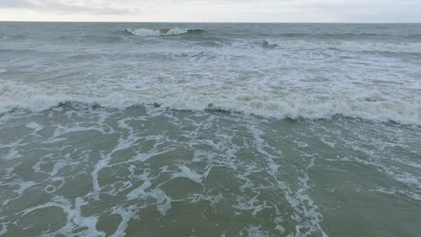 aerial view of big stormy waves breaking against the white sand beach, overcast day, seashore dunes damaged by waves, coastal erosion, climate changes, wide low drone shot moving forward