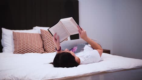 a young woman is reading a book with black hardcover in her bedroom, laying on the bed, she spends her free time with a book, relaxing at home, turns the page, flips the page, close-up shot