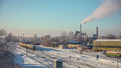 Der-Industrielle-Teil-Einer-Stadt-Im-Winter-Mit-Einem-Vorbeifahrenden-Kesselwagenzug,-Während-Dampf-Aus-Einem-Fabrikschornstein-Kommt---Zeitraffer