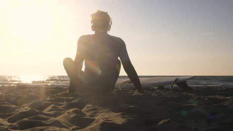 joven surfista en traje de buceo sentado con una tabla de surf acostado en la arena en la playa escuchando música bailando mirando el océano al atardecer
