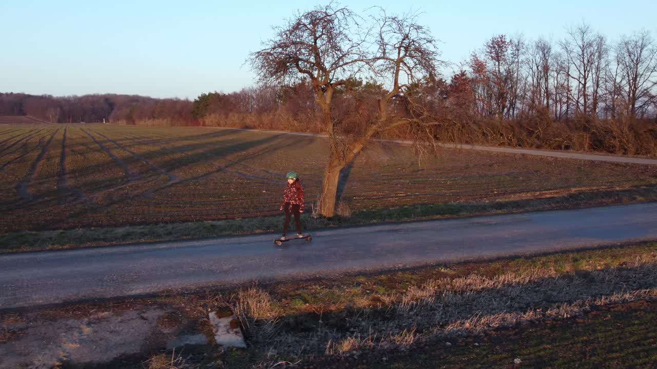 girl riding an electric longboard on a road surrounded by trees, lit by sunset in spring, aerial