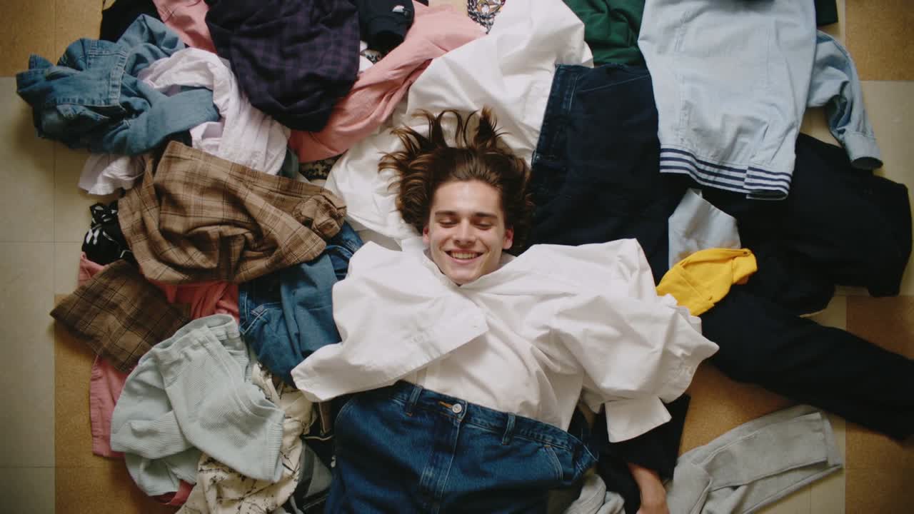 Holding top cam filming teen lying on clothes pile in bedroom, wearing white shirt, jeans, resting