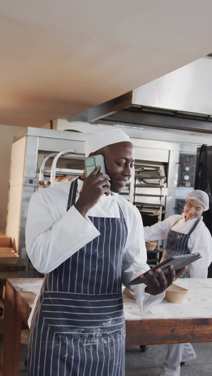 Vertical video of african american male baker talking on smartphone in bakery kitchen, slow motion