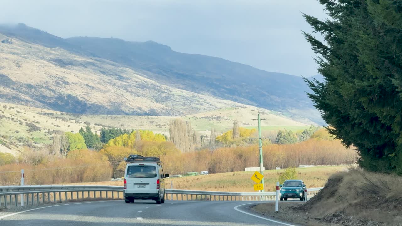 A vehicle travels along a winding road in Wanaka, New Zealand, surrounded by mountains and trees under clear daylight