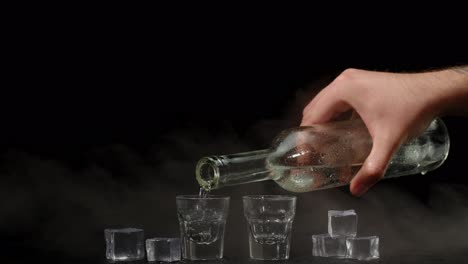 Bartender-pouring-up-two-shots-of-vodka-with-ice-cubes-from-bottle-into-glasses-on-black-background