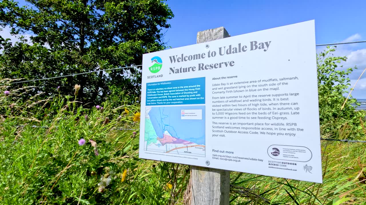 Camera slowly pans past an informational sign for Udale Bay Nature Reserve, surrounded by lush greenery and bright daylight in Cromarty, Scotland