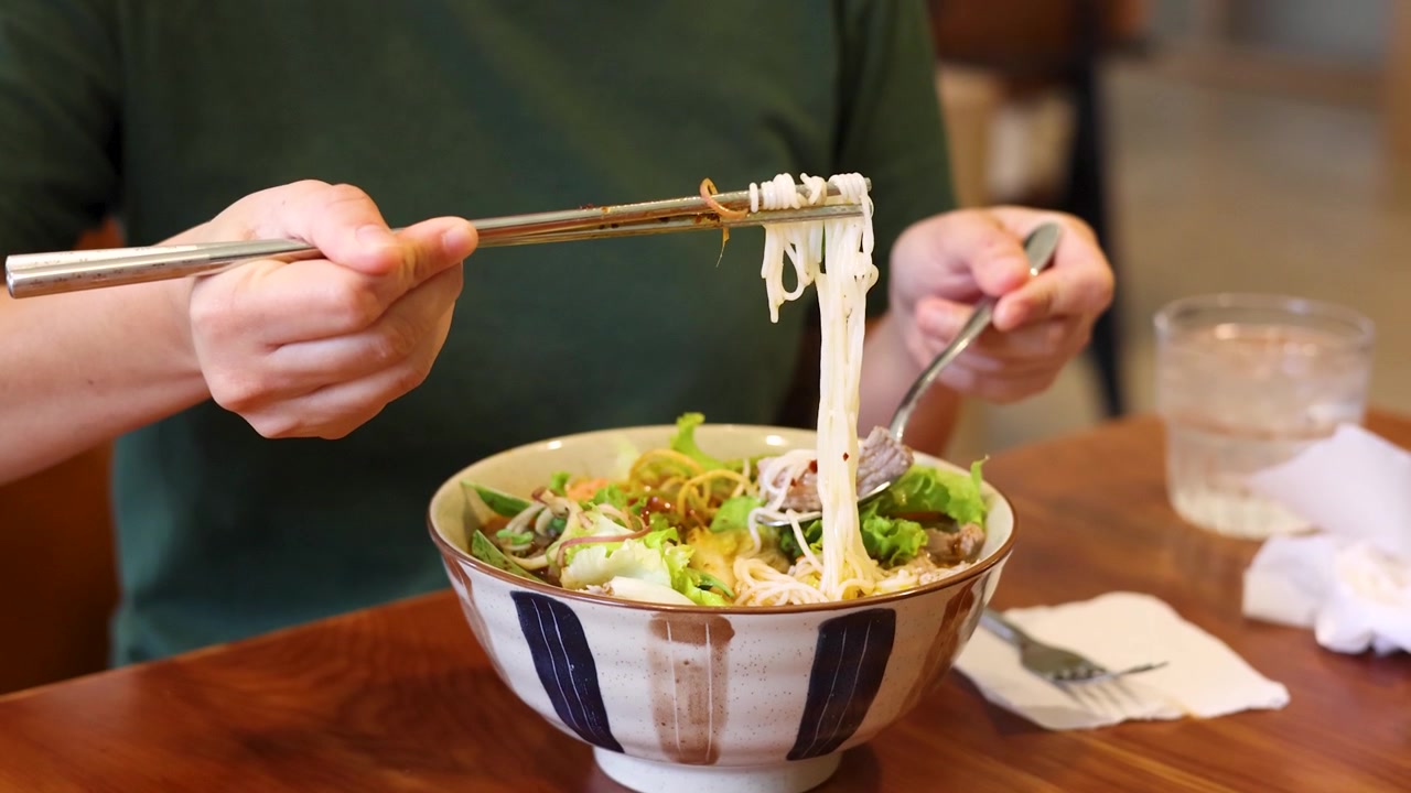 Una persona usa le bacchette per sollevare spaghetti di riso da una ciotola di ceramica in un ristorante