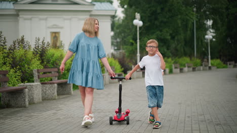 a mother walks alongside her son, who holds onto a scooter, in the background, the younger child runs happily to catch up to them as he hold his mum hand