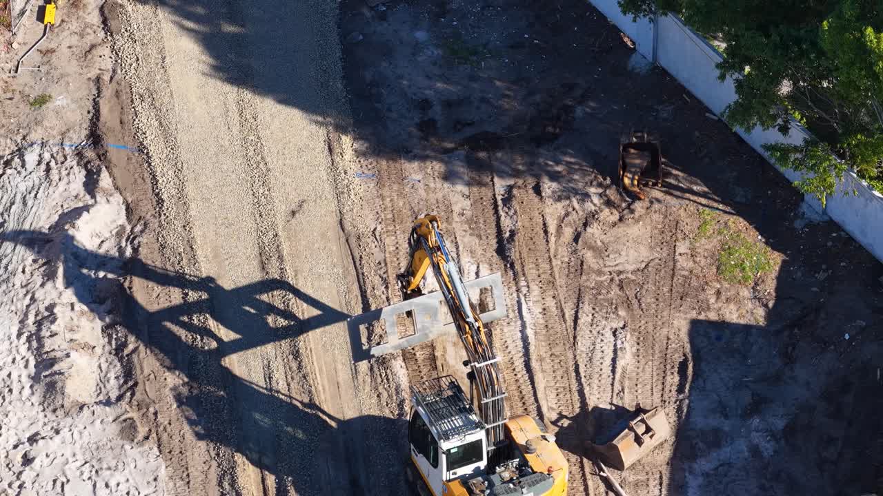 Excavator moves soil on dirt road at construction site, captured in bright daylight aerial view
