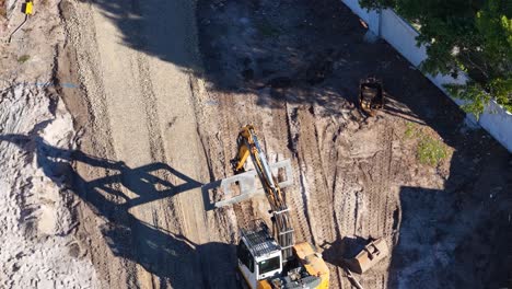 excavator moves soil on dirt road at construction site, captured in bright daylight aerial view