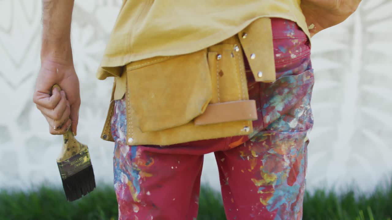 video de la sección media del artista masculino caucásico con un pincel de pie frente al mural en la pared
