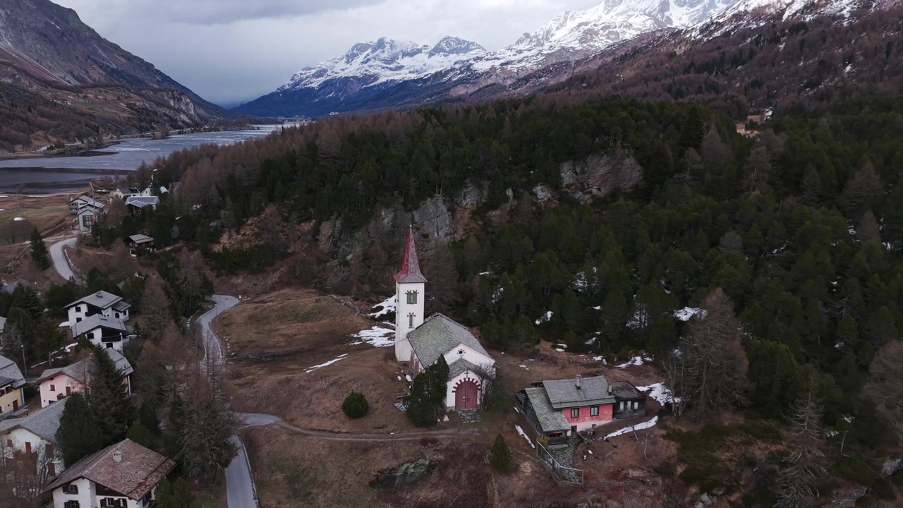 Drone uzaktaki karlı zirvelere doğru uzaklaşırken, Maloja köyünün üzerinde kırmızı kuleli küçük bir kilise duruyor.