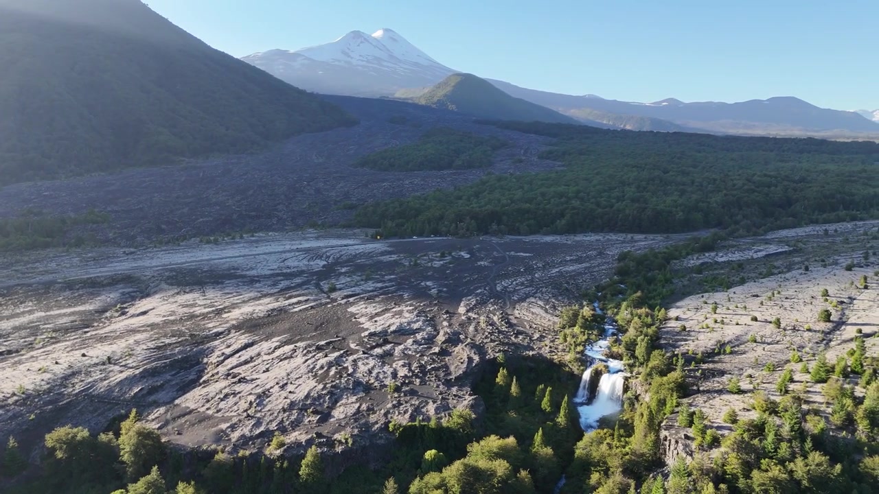 Aerial drone footage of Truful Truful River waterfall cascading through dense forest surrounded by vast ancient lava fields from Llaima volcano. Clear sunny day with snowcapped peak visible