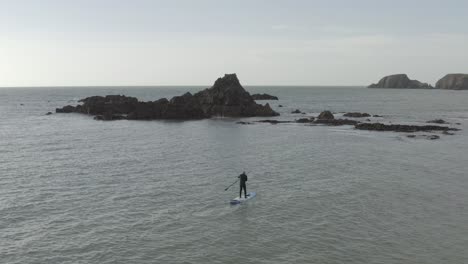 stand up paddleboarder paddles toward rocky ocean islets on grey day