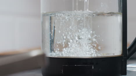 primer plano de un frasco de agua eléctrica con agua que se vierte en él, con burbujas de agua que se forman y una inscripción en el frasco, en un entorno de cocina limpio con electrodomésticos modernos