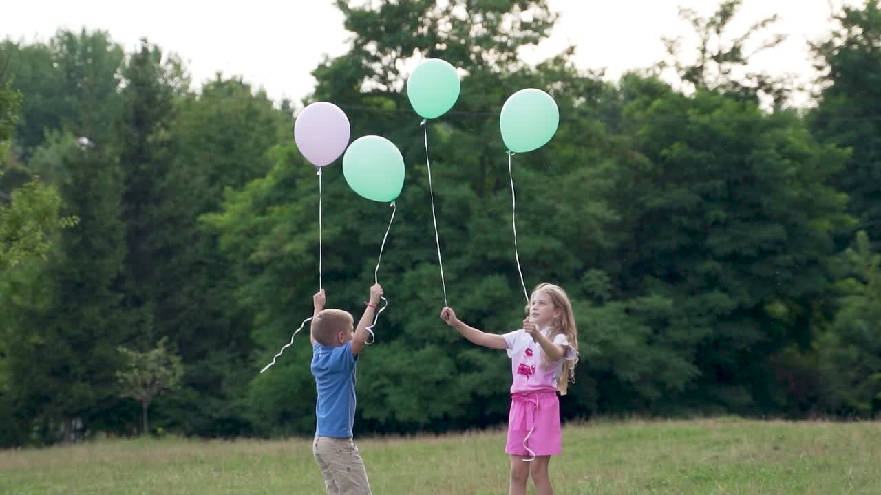 Joyful boy and girl are playing with balloons in the park with trees. Children's holiday. A girl in pink is jumping on the lawn