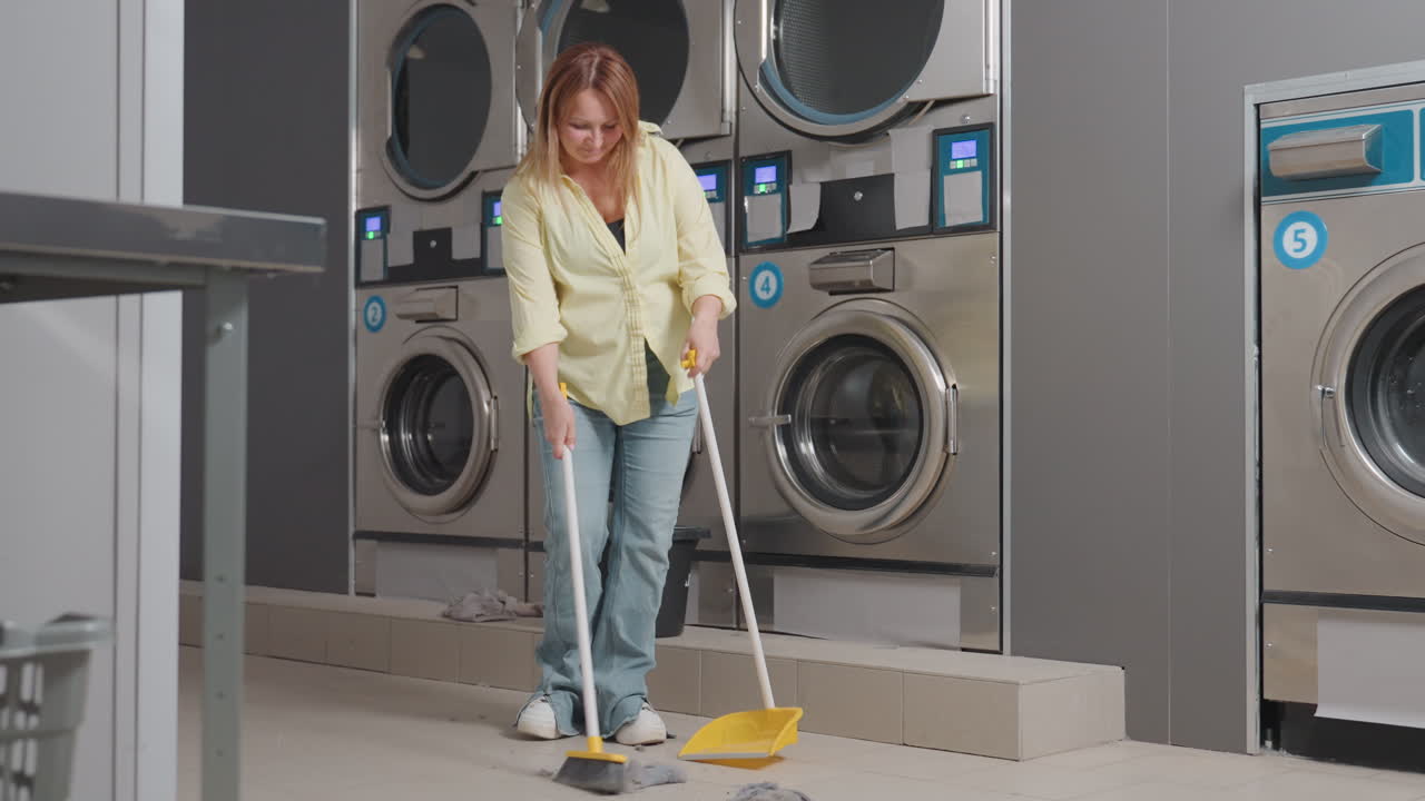 Excited work woman dances with broom stick and dust pan near industrial washer in laundromat, cleaning lint spilled from filter drawer on floor, smiling caretaker enjoying tidy workspace maintenance