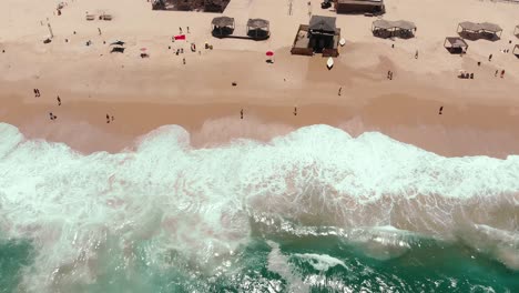 Static-aerial-drone-showing-endless-wild-sea-waves-breaking-on-the-sandy-coast-line-while-people-hang-out-on-the-beach-and-a-life-guard-station-is-in-the-middle