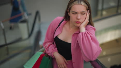 lady in pink outfit, resting her chin on her hand, holding a shopping bag in her right hand, gazes thoughtfully while riding on a moving escalator, with blurred view of people in the background