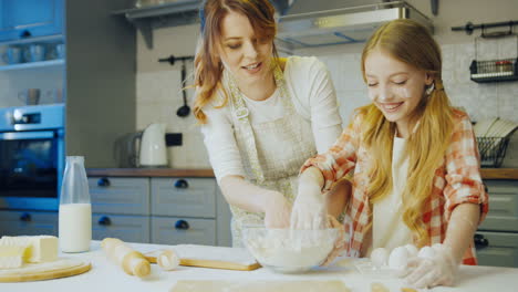 Pretty-mother-showing-her-little-blonde-daughter-how-to-knead-a-flour-and-she-trying-to-do-this-on-the-table-in-the-nice-kitchen.-Inside