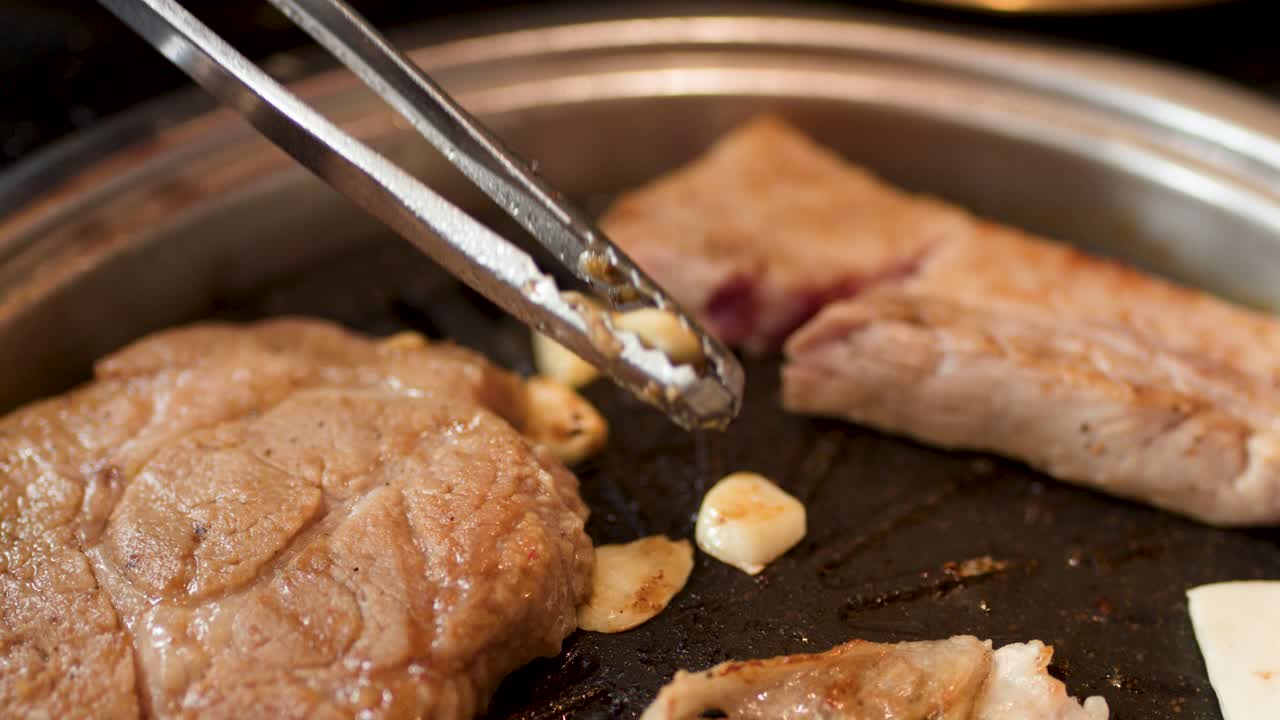 Metal tongs flip garlic cloves on a sizzling Korean barbecue grill with pork and tofu