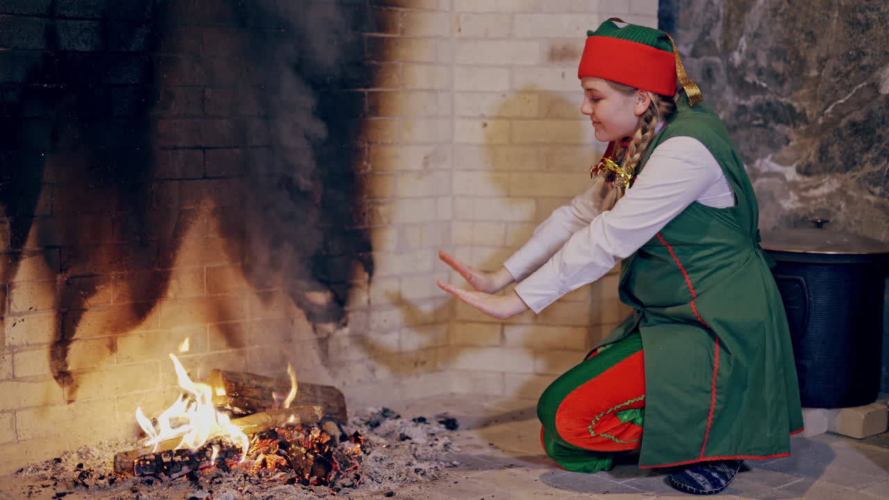 Beautiful elf is getting warm while sitting near the fire at fireplace. Female elf in fairy costume rubs her cold hands close to the fireside. Christmas atmosphere indoors.