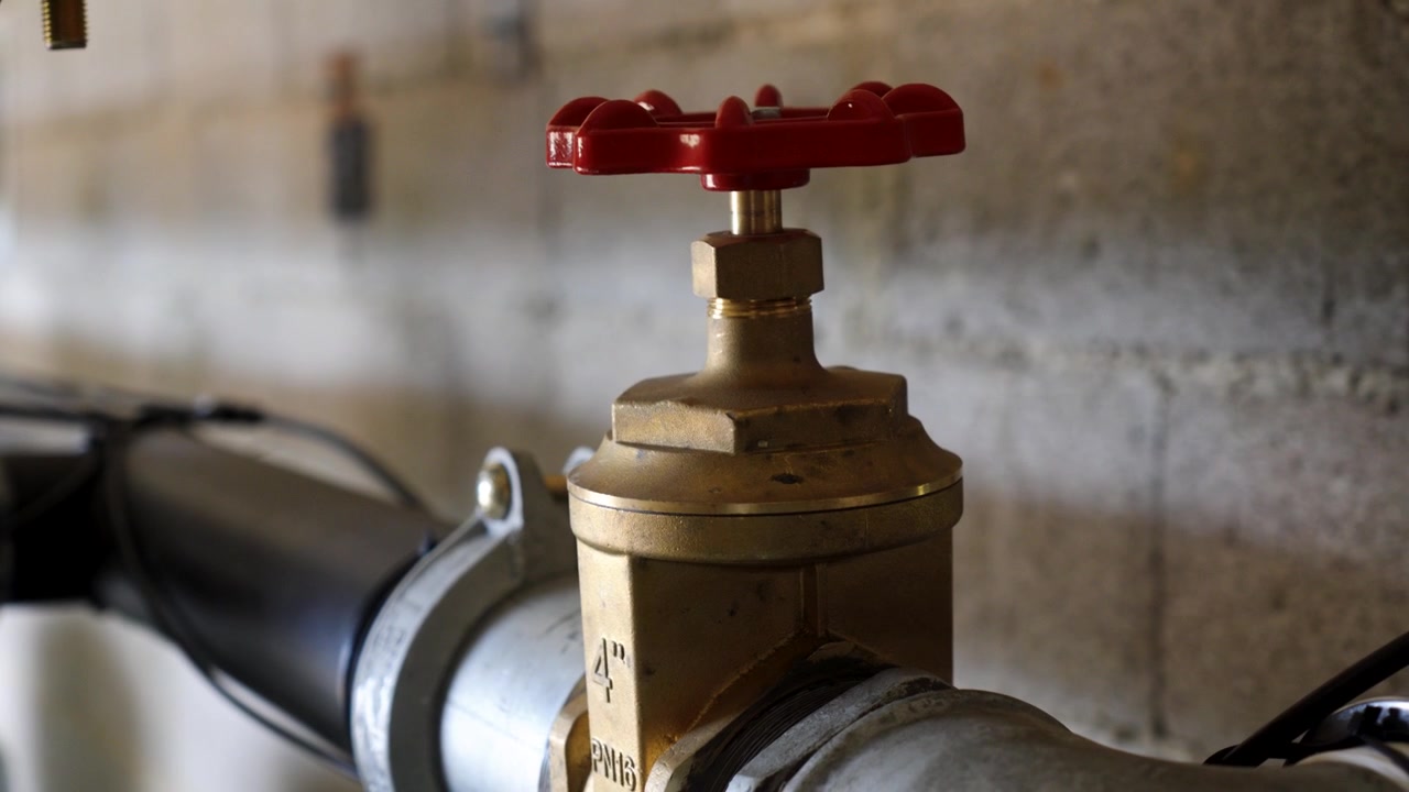 close-up of a smart new valve fitted between two pipes, with a red wheel on top and a gold body, grey pipes and a breeze block wall in the background