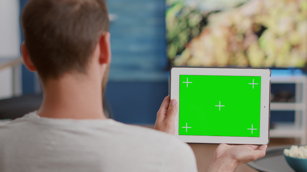 Closeup of man holding digital tablet with green screen watching online video content sitting on sofa
