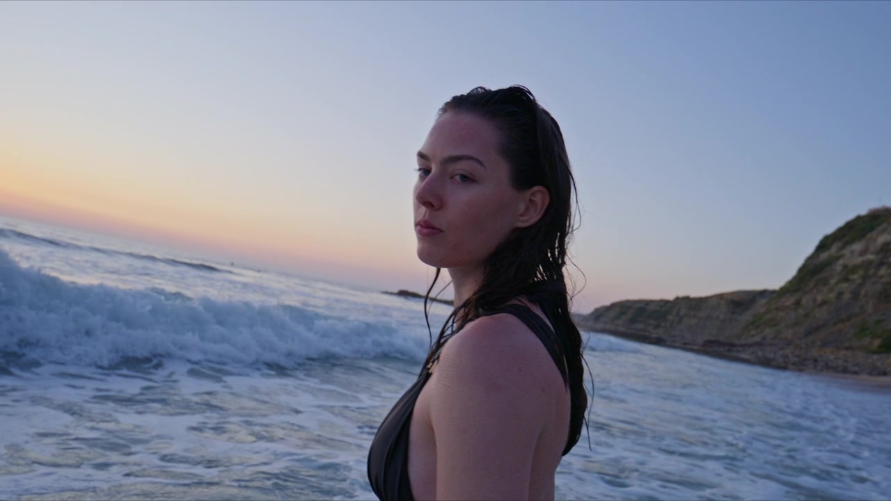 Close up portrait of a woman with wet hair standing by ocean waves at sunset, looking at the camera with a calm cinematic beach fashion mood.