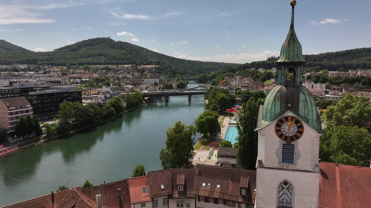 clocktower rises over rooftops as river curves toward olten cityscape under summer sky