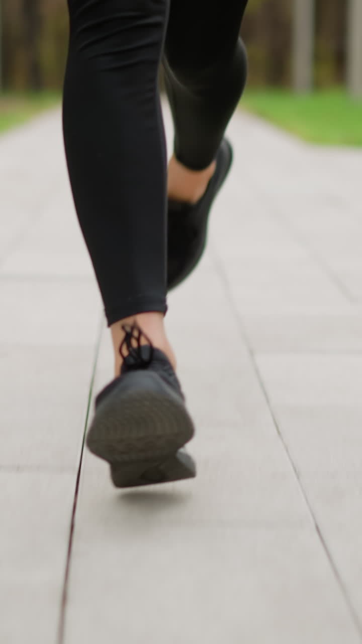 Close-up leg view of runner wearing black sneakers running on a paved track through park, blurred background of green grass, highlighting athletic movement and jogging activity