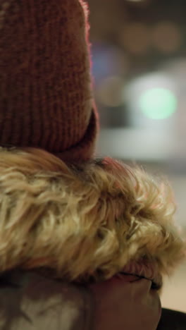 first-person view of a woman in a winter coat with a fur hood, standing still at night with blurry moving cars and colorful lights in the background