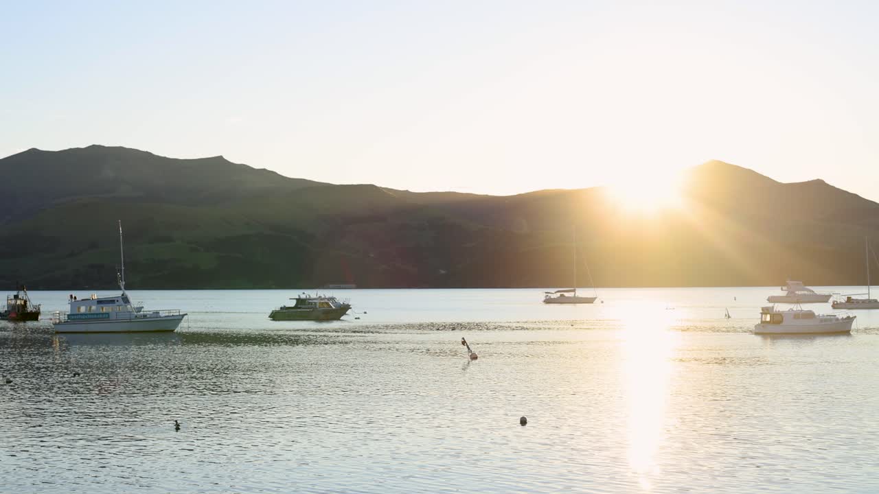 Boats gently float in Akaroa Harbor as the sun sets behind the mountains, casting golden rays over the tranquil water