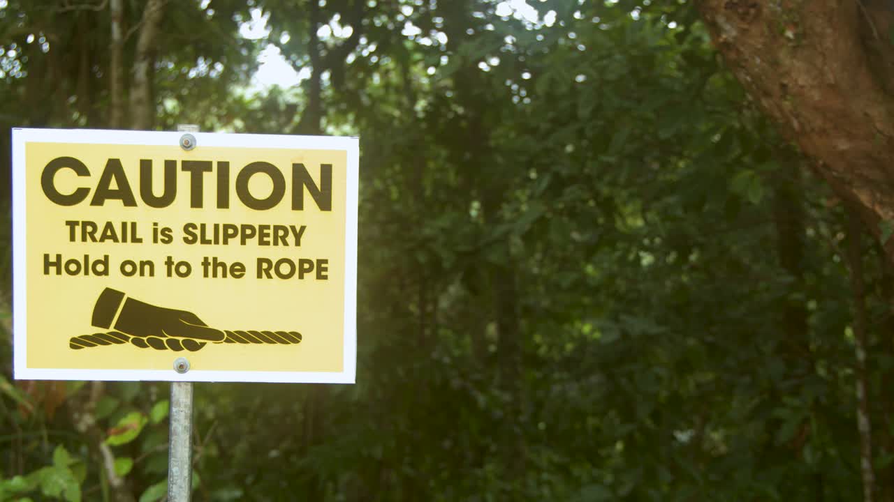 Ein gelbes Warnschild warnt Wanderer vor rutschigen Wegen in einer üppigen tropischen Waldumgebung.