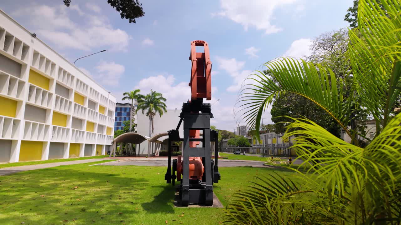 A construction drill is positioned centrally in a vibrant campus setting, with modern buildings and lush greenery surrounding it. The camera zooms out to capture the entire scene in bright daylight