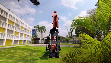 a construction drill is positioned centrally in a vibrant campus setting, with modern buildings and lush greenery surrounding it. the camera zooms out to capture the entire scene in bright daylight