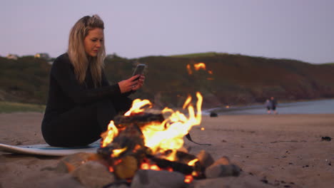 woman sitting on surfboard by camp fire on beach using mobile phone as sun sets behind her