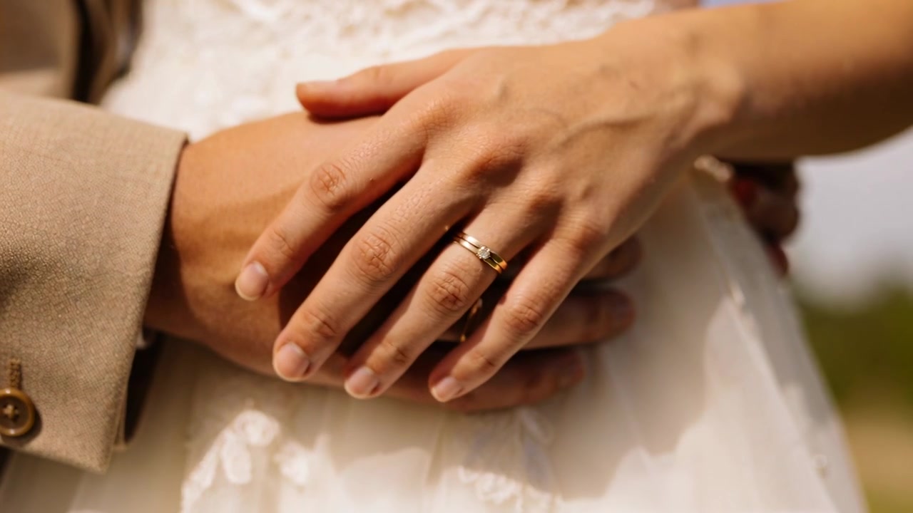 Anelli scintillanti, sposa in pizzo che posa le dita sulle mani dello sposo, abito beige, giardino per matrimonio.