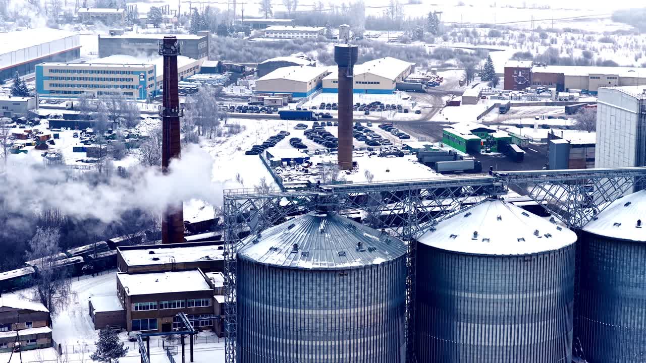 Dynamische Luftbild-Parallaxe, die hohe Aufzugssilos, Schornsteine und Industriegebäude umkreist, die mit Schnee bedeckt sind, wobei Dampf in Jelgava, Lettland, aufsteigt