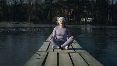 saque el zoom de la mujer caucásica sentada en el muelle y meditando con los ojos cerrados en el invierno.