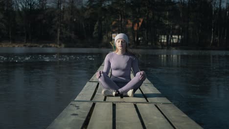 zoom out of caucasian woman sitting on pier and meditating with eyes closed in the winter.