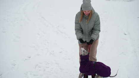 dog wearing purple coat stands on hind legs reaching toward woman in winter outfit during outdoor training session on snow-covered ground, red leash dangling as playful interaction unfolds in cold park