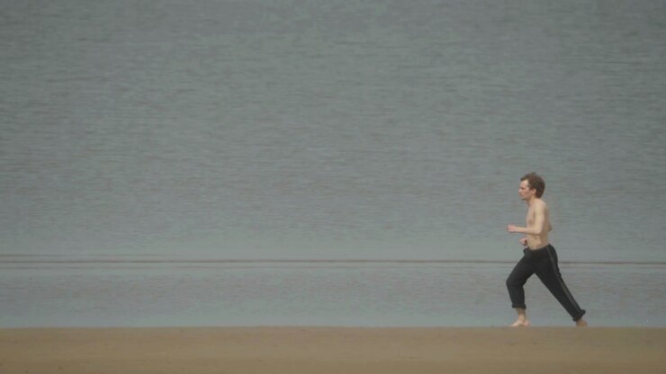 Young shirtless adult male at water’s edge on a beach, slow motion