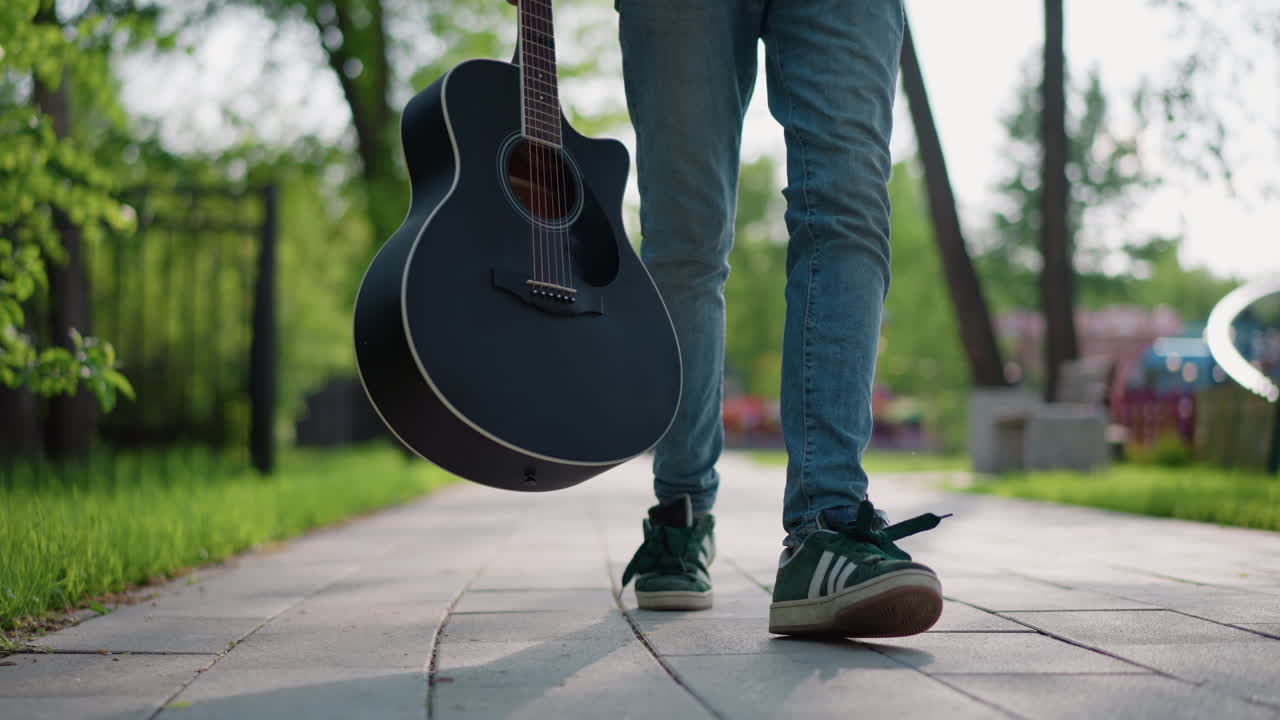 Güneşli park yolunda yürüyen gitarist, kot pantolon ve spor ayakkabıları görülebiliyor, siyah akustik gitar yanında sallanıyor, kaldırımda rahat adımlarla yürüyor, rahat banliyö yeşilliği ve sabah ışığı atmosferi yansıtıyor