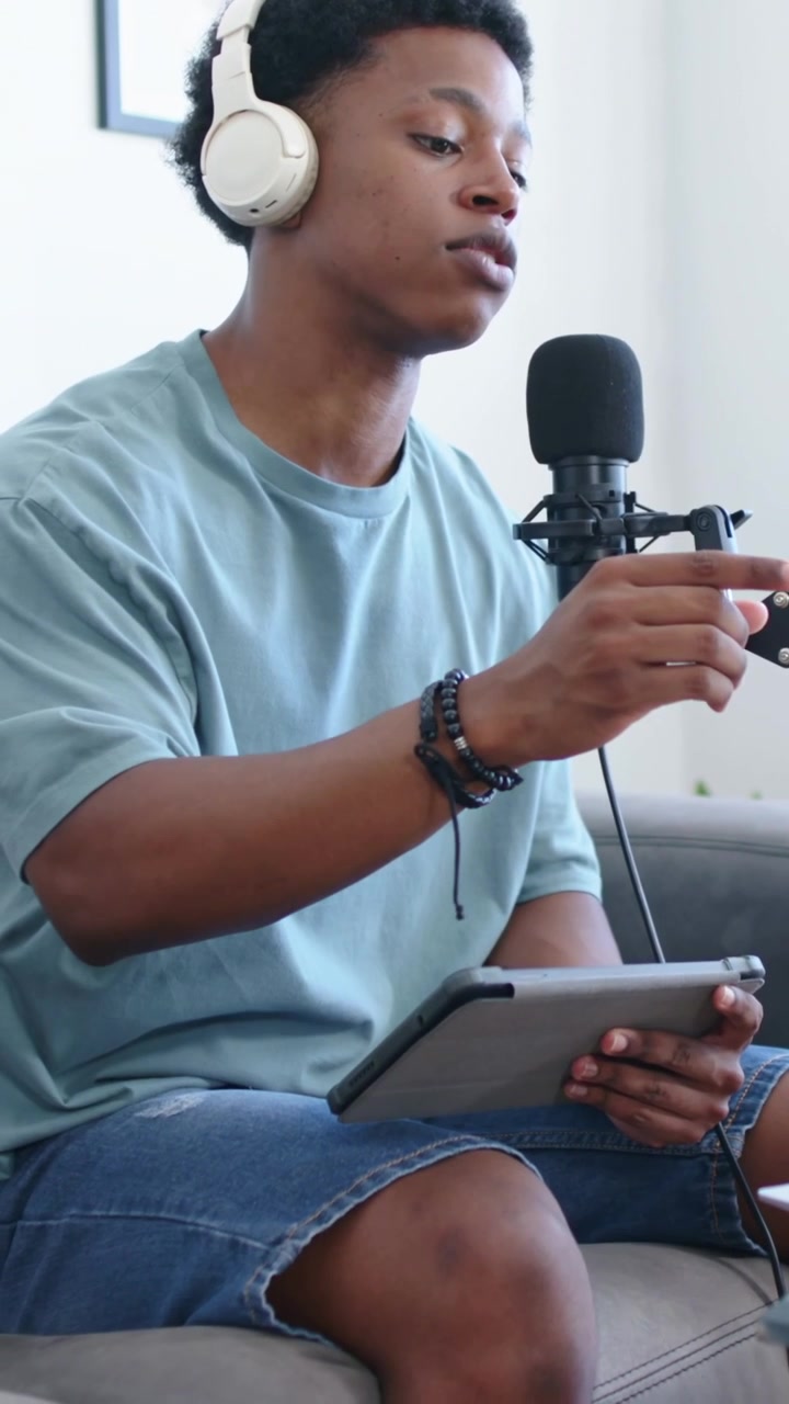 Vertical video: Glancing at mic, host with white headphones reading tablet recording in home studio