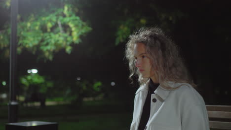 student sitting alone outdoors at night lost in thought after reading book, with dim park lights glowing in blurred background and soft breeze gently blowing through hair